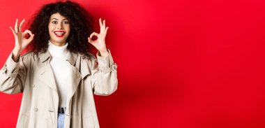 Trençkotlu beyaz kadın hoş bir jest yapıyor ve gülümsüyor. Kırmızı arka planda duran bir arkadaş öneriyor..