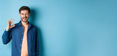 Olumlu gülümseyen adam göz kırpıyor ve iyiye işaret gösteriyor, iyi işi övüyor, iyi bir arkadaş tavsiye ediyor, mavi arka planda duruyor..