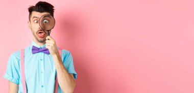 Papyonlu komik adam büyüteçten bakıyor, şaşı bakıyor ve pembe arka planda aptal suratlar yapıyor..