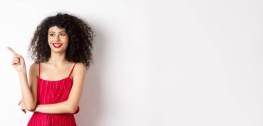 Kırmızı elbiseli ve akşam makyajlı zarif bir kız, solu işaret ediyor ve logoya bakıyor, tatmin olmuş gülümsüyor, beyaz arka planda duruyor..