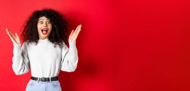 Makyajlı ve tişörtlü mutlu ve şaşırmış kıvırcık bir kadın resmi. İyi haberlerle el kaldırıyor ve seviniyor. Kırmızı arka planda duruyor..