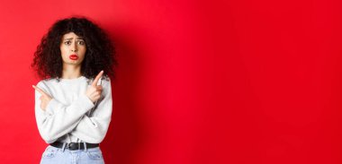 Kıvırcık saçlı gergin üzgün kız, hıçkıra hıçkıra ağlıyor ve parmaklarını yana doğru gösteriyor, iki şey gösteriyor ve kararsız, kırmızı arka plan.