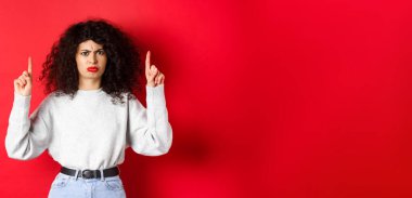 Huysuz, kıvırcık saçlı, kaşlarını çatmış, tatminsiz bir şekilde yüzünü ekşiten, kötü bir şeyi işaret eden, şirketten şikayet eden, kırmızı arka planlı genç bir kadın..