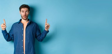 Şaşırmış genç adam diyor ki: Vay canına, reklamı kontrol ediyor, parmaklarını havaya kaldırıyor ve etkilenmiş görünüyor, mavi arka planda duruyor..
