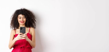 Mutlu modaya uygun bir kadın akıllı telefonda fotoğraf çekiyor, cep telefonunda fotoğraf çekiyor ve gülümsüyor, beyaz arka planda kırmızı elbiseyle dikiliyor..