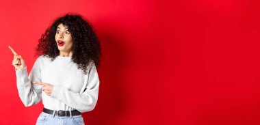 Şaşırmış kıvırcık saçlı, boş alanı gösteren, logo ya da reklam veren, kırmızı arka planda duran, kıvırcık saçlı, çekici kız..