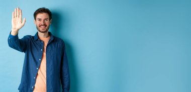 Dost canlısı genç adam gülümsüyor ve beşlik çakıyor, selam vermek için feragat ediyor, mavi arka planda duruyor..