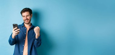 Şanslı adamın çevrimiçi kazandığı, cep telefonu ekranını okuduğu ve yumruk pompası yaptığı, zafer kazandığı, mavi arka planda durduğu görüntü.