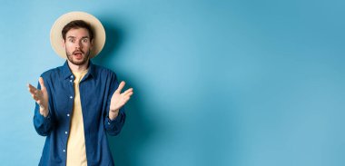 Şok olmuş erkek turist el kaldırıyor ve kameraya bakıyor. İnançsızlıkla büyük habere tepki veriyor. Mavi arka planda saman şapka ve tişörtle duruyor..