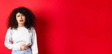 Kadın hasta, eğiliyor ve karnına dokunuyor, karın ağrısı ya da regl sancısı çekiyor, kırmızı arka planda duruyor..