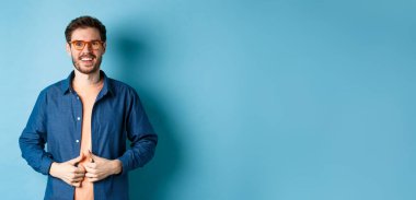 Yakışıklı sakallı adam gülümsüyor, yeni gözlük takıyor, mavi arka planda duruyor..