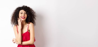 Parti elbisesi içinde çekici bir kadın, gülüyor ve eğleniyor, kameraya mutlu gülümsüyor, makyaj kırmızı ruj sürüyor, beyaz arka planda duruyor..