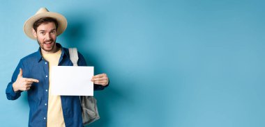 Yakışıklı turist gülümsüyor ve boş kağıt parçasına işaret ediyor, hasır şapka ve sırt çantası takıyor. Yaz tatili kavramı.