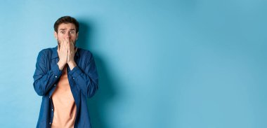 Korkmuş endişeli adam elleriyle ağzını kapatıyor ve mavi arka planda duran korkunç bir şeye bakıyor..