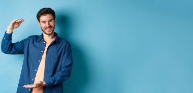 Yakışıklı beyaz adam büyük bir şey gösteriyor, büyük bir nesne şekillendiriyor, elleri ve gülüyor, mavi arka planda duruyor..