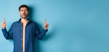Kafası karışmış genç adam kaşlarını çatıyor, parmaklarını havaya kaldırıyor ve hoşnutsuz görünüyor, mavi arka planda duruyor. Boşluğu kopyala