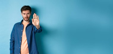 Üzgün adam kaşlarını çatıyor ve durmak istiyor, yasaklamak için elini uzatıyor ya da kötü bir şeye karşı çıkıyor, mavi arka planda duruyor..