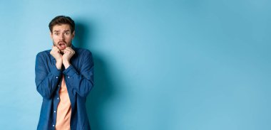 Korkmuş genç adam nefes nefese ve çığlık atıyor korkmuş, bir şeye bakıyor, mavi arka planda duruyor..