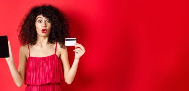 İnternetten alışveriş konsepti. Kırmızı elbiseli ve rujlu güzel bir kadın, boş telefon ekranı ve plastik kredi kartı gösteriyor, stüdyo arka planında duruyor..