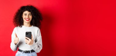 Gülümseyen Avrupalı kadın mutlu ve memnun görünüyor, cep telefonu kullanıyor ve başparmak yapıyor, başvuru öneriyor, kırmızı arka planda duruyor..