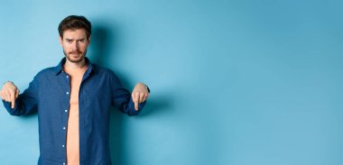 Endişeli genç adam, somurtuyor ve endişeli görünüyor, parmakları aşağıda, kötü bir şey gösteriyor, mavi arka planda duruyor..
