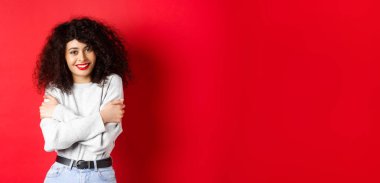 Şefkatli genç bir kadın kendini kucaklıyor, rahat ve mutlu hissediyor, kameraya aptalca gülümsüyor, kırmızı arka planda duruyor..