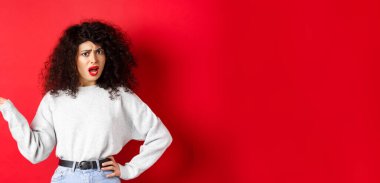 Sinirli ve kafası karışık, kıvırcık saçlı beyaz kadın, elini kaldırıyor ve tartışıyor, tartışıyor, bir şey anlayamıyor, kırmızı arka planda duruyor..