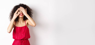 Kıvırcık saçlı, kırmızı elbiseli, elleriyle gözlerini kapatan ama parmaklarının arasından bakıp gülümseyen, ilginç, beyaz bir arka plana bakan komik bir genç kadın..