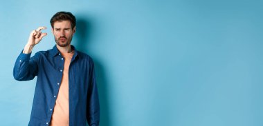 Kafası karışmış bir adam, küçük boyda, şüpheli ya da şüpheci görünüyor, mavi arka planda duruyor. Boşluğu kopyala