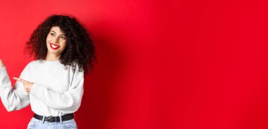 Kıvırcık saçlı ve makyajlı güzel bir Avrupalı kadın, parmaklarını sola uzatıyor ve rüya gibi bir gülümsemeyle yan yana bakıyor, terfi anlaşmasına bakıyor, kırmızı arka plan..