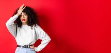 Sinirli ve yorgun genç bir kadın alnını tokatlıyor, facepalm yapıyor ve kameraya bakıyor şaşkın şaşkın, kırmızı arka planda duruyor..