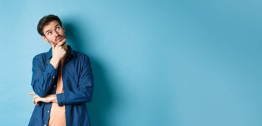 Düşünceli genç adam yüzüne dokunuyor ve boş alana bakıyor, bir şeyler hayal ediyor, plan dışı düşünüyor, mavi arka planda duruyor..