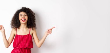 Kırmızı elbiseli mutlu güzel bayan, gülüyor ve yandan işaret ediyor, iki yol gösteriyor, beyaz arka planda duruyor..