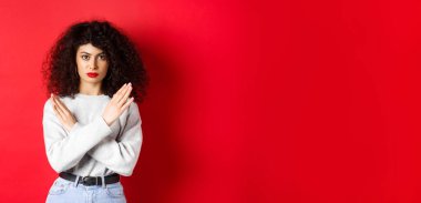 Kıvırcık saçlı, ciddi bir beyaz kadın hayır diyor. Kırmızı arka planda hoşnutsuz duran, kötü bir şeyi engellemek için çapraz jest yapıyor..