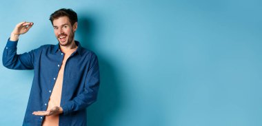 Neşeli modern adam gülümsüyor, büyük boy gösteriyor ve mutlu görünüyor, mavi arka planda duruyor. Boşluğu kopyala