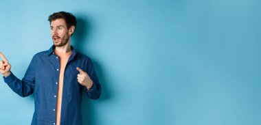 Kafkasyalı beyaz adam solu işaret ediyor, reklamlara bakıyor, mavi arka planda duruyor..
