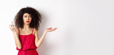 Kıvırcık saçlı üzgün kız arkadaş, yüzük takmayan parmağını gösteriyor ve kafası karışmış görünüyor, kararsız erkek arkadaşından şikayetçi, evlenmek istiyor, beyaz arka planda duruyor..