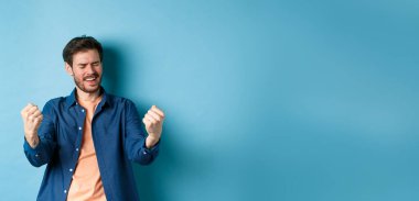 Başarıyı kutlayan mutlu bir adam tasviri, evet diye bağırıyor ve yumruklarını sallıyor, hedefe ulaşıyor ve başarıyor, kazanıyor ve dans ediyor, mavi arka planda duruyor..