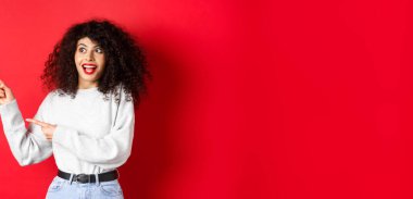 Heyecanlı, kıvırcık saçlı ve kırmızı dudaklı güzel kız, parmaklarını şaşkın bir şekilde sola çeviriyor, afişi gösteriyor, stüdyo arka planında duruyor..
