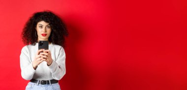 Kıvırcık saçlı, akıllı telefonuyla video kaydeden, cep telefonuyla fotoğraf çeken ve kırmızı arka planda duran genç bir kadın..