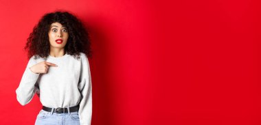 Kendini gösteren, kaşlarını kaldıran ve kameraya inanmayan, kırmızı arka planda duran çekici bir manken..