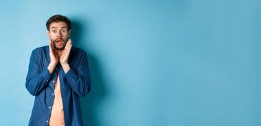 Şok olmuş genç adam ürkmüş görünüyor, endişeyle bağırıyor, mavi arka planda duruyor. Boşluğu kopyala