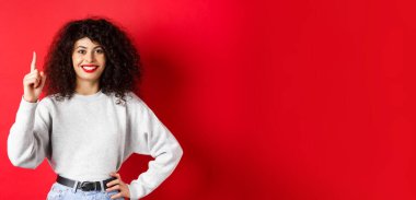 Bir numarayı gösteren kıvırcık saçlı modern Avrupalı kadın portresi, sipariş veriyor, parmağını kaldırıyor ve gülümsüyor, kırmızı arka planda duruyor..