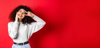 Güzellik ve makyaj. Kıvırcık saçlı mutlu bir genç kadın, dokunaklı bir yüz ve sevimli bir gülümsemeyle V işareti gösteriyor, kırmızı arka planda duruyor..