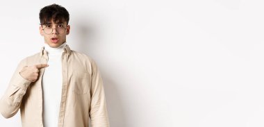 Kafası karışmış ve şaşırmış genç bir adam kendini işaret ediyor, seçiliyor ya da suçlanıyor, beyaz arka planda duruyor..