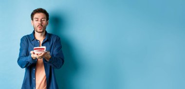 Kutlama ve tatil konsepti. Yakışıklı genç adam doğum günü pastasında mum üflüyor ve dilek diliyor, mavi arka planda duruyor..