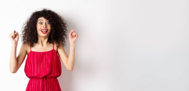 Heyecanlı, kıvırcık saçlı, akşam makyajlı, nefes nefese, el kaldıran ve sevinen, ödül kazanan ve kutlayan beyaz arka planda duran bir kadın..