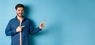 Sakallı neşeli beyaz adam boş bir yeri işaret ediyor ve kameraya gülümsüyor. Reklam yapıyor, mavi arka plan..