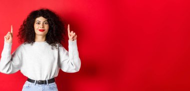 Kıvırcık saç stili ve kırmızı dudaklı neşeli bir kadın resmi. Boş yerleri işaret ediyor, reklam yapıyor, stüdyo arka planında günlük giysiler içinde duruyor..