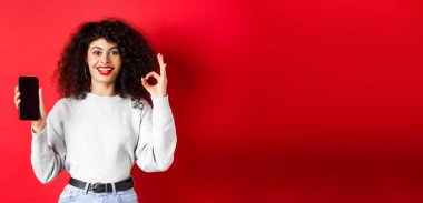 Kıvırcık saçlı neşeli Avrupalı kadın, boş cep telefonu ekranı gösteriyor ve tamam jest, gülümsüyor tatmin olmuş, iyi bir uygulama ya da terfi, kırmızı arka plan.